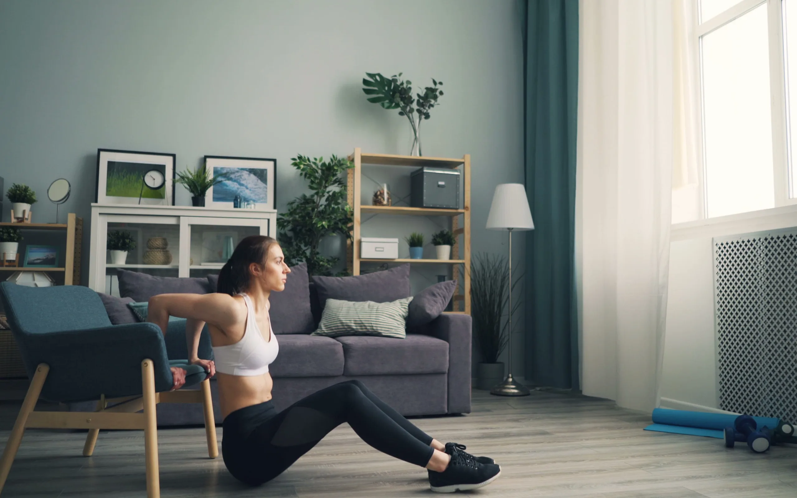Une personne en tenue de sport fait des dips sur une chaise, dans un salon clair.